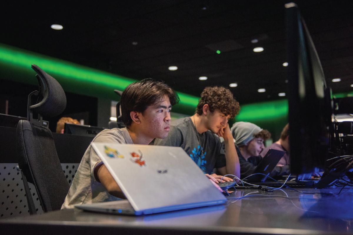 Participants work on laptops and monitors in a cybersecurity lab during a hackathon, collaborating at computer stations under green accent lighting.