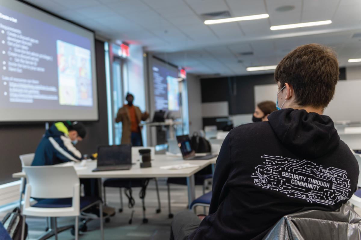 Participant wearing a RITSEC hoodie sits in a classroom during a cybersecurity workshop while a presenter speaks near projected slides and attendees work at tables.