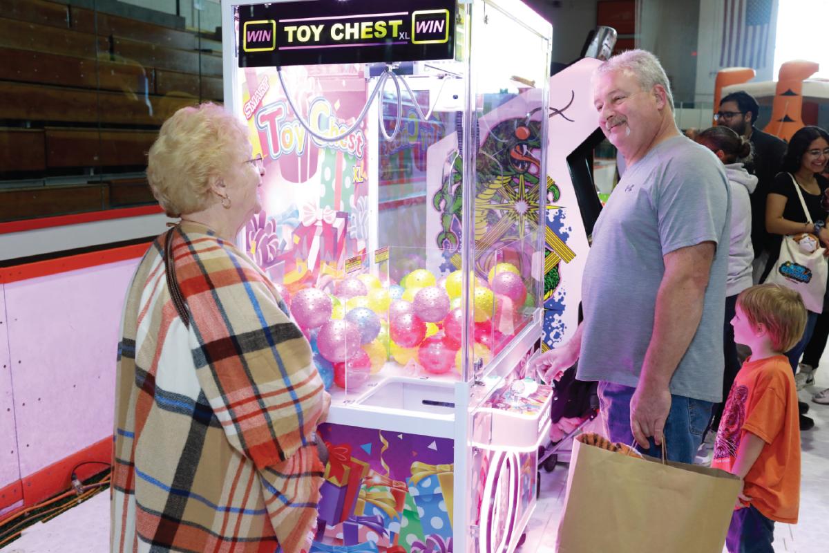 Participants and families take turns playing a claw machine game in a busy indoor Family Fun Zone surrounded by arcade attractions and activities.