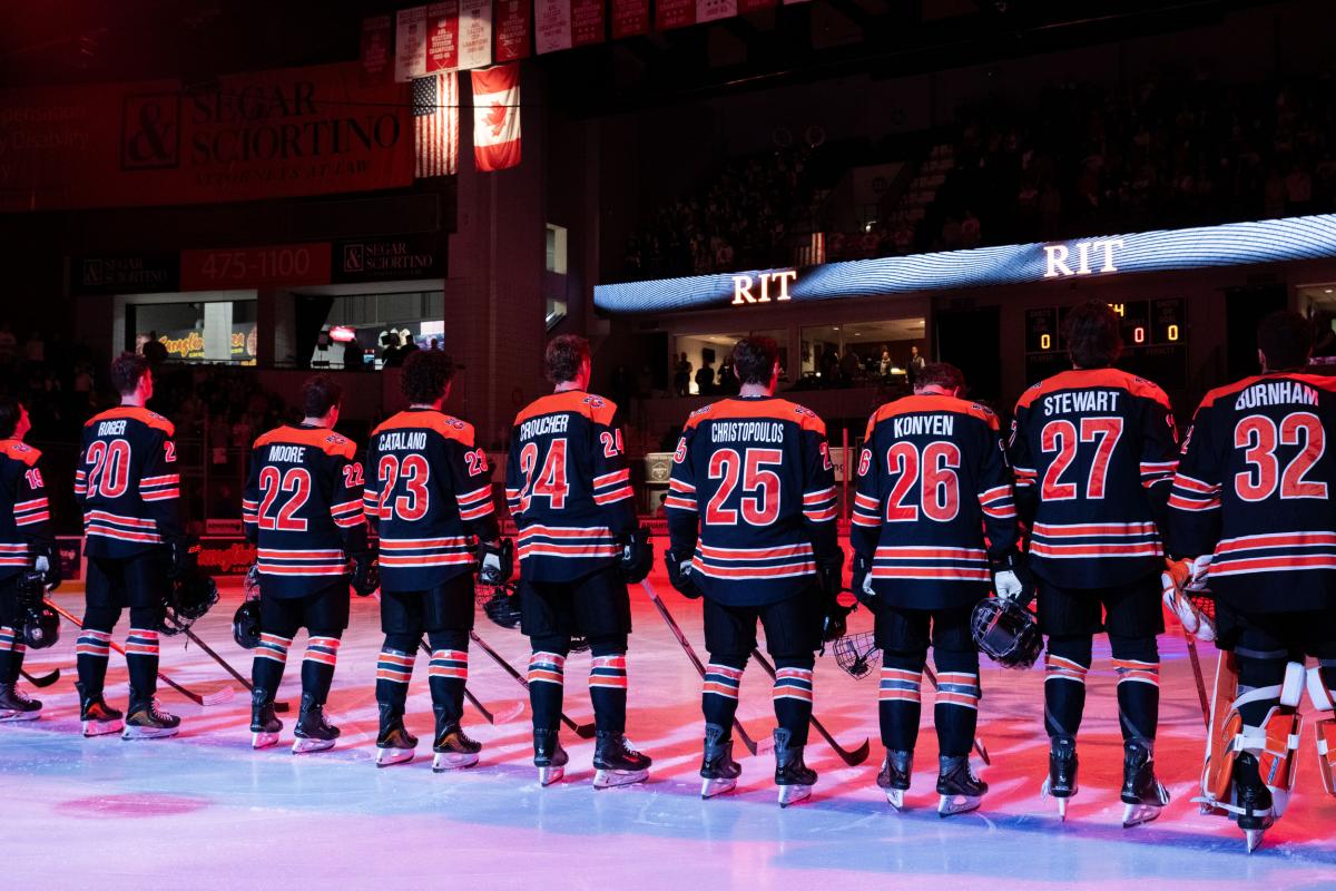 RIT hockey players line up on the ice during pregame introductions, with fans filling the arena stands in the background.