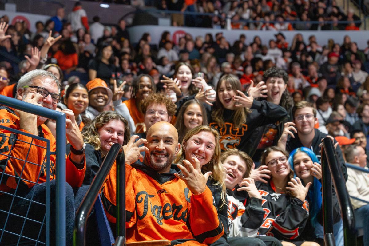 RIT hockey fans wearing team apparel sit together in a crowded arena during a game.