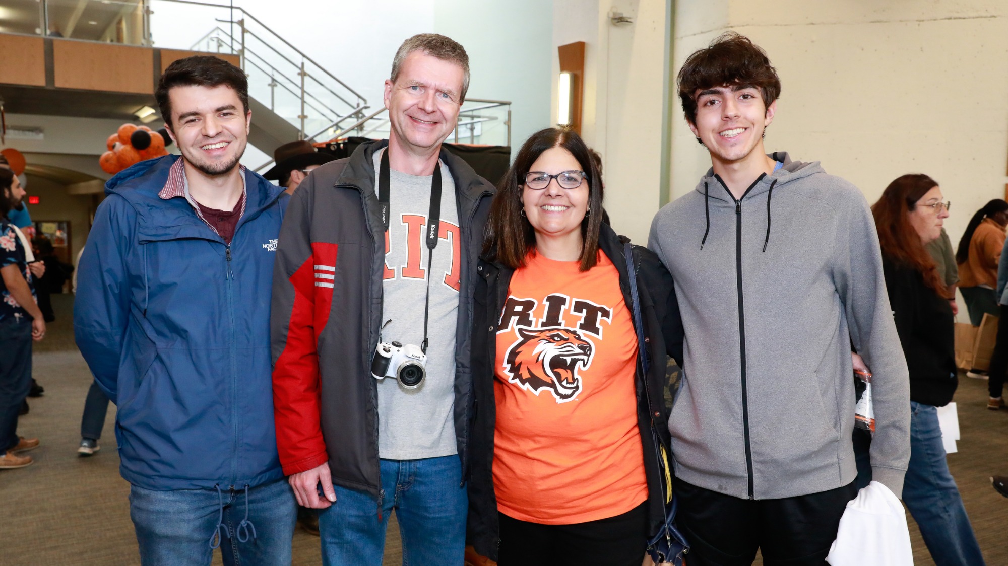 Four visitors stand together for a photo in the RIT Welcome Center during an event, with other attendees visible in the background.