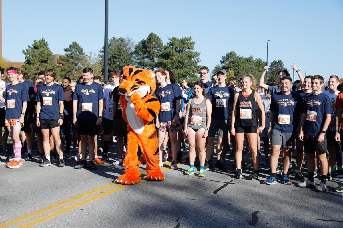 Runners gather at the starting line of an RIT 5K race, with RITchie the Tiger standing among participants.