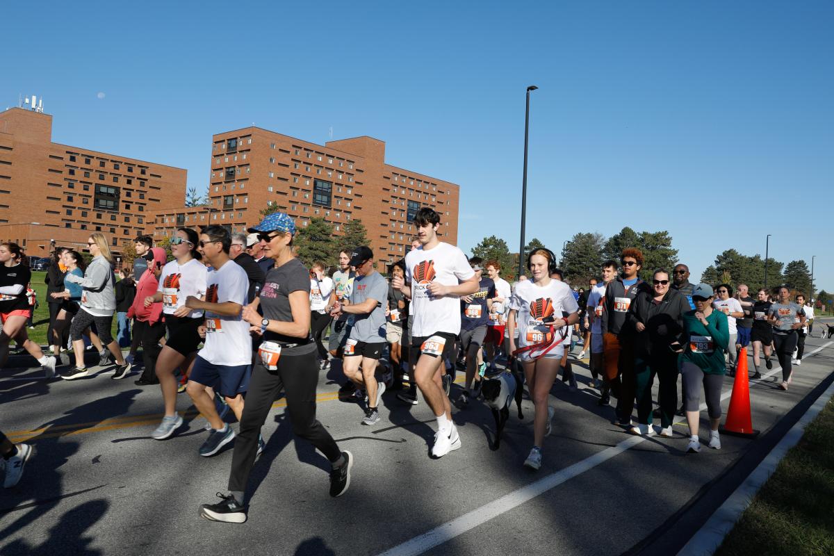 Participants run and walk along a campus roadway during an RIT 5K event, with residence halls visible in the background.