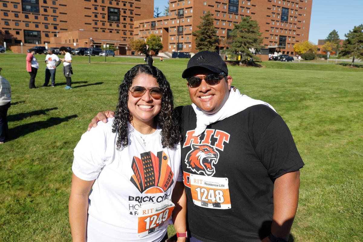 Two participants wearing race bibs stand together on a grassy area during an RIT 5K event, with campus residence halls in the background.