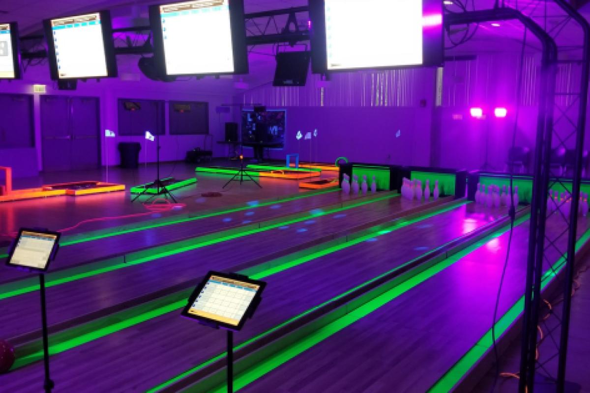 Indoor bowling alley featuring glowing lanes, illuminated pins, and overhead scoring screens under colorful neon lighting.