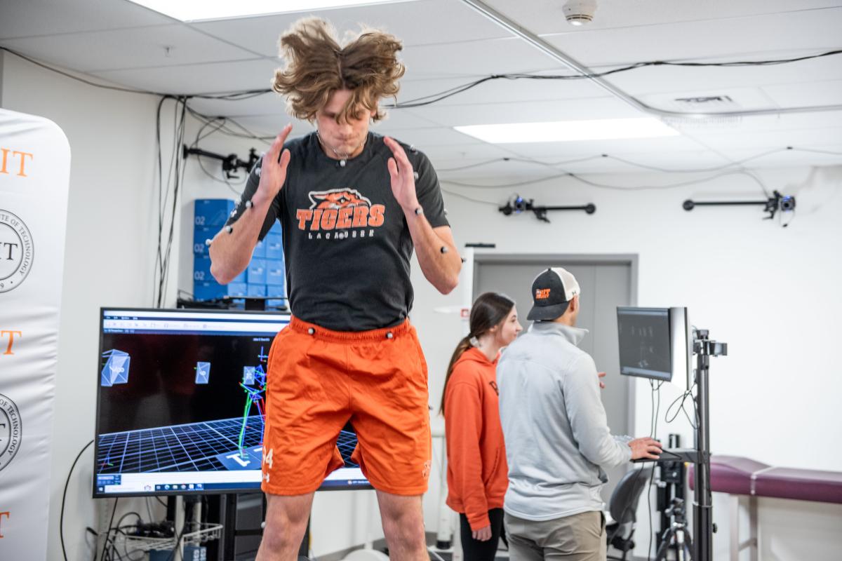 Student athlete wearing an RIT Tigers Lacrosse shirt and motion-capture sensors jumps in a human movement laboratory while researchers monitor data on computer screens and cameras track movement.