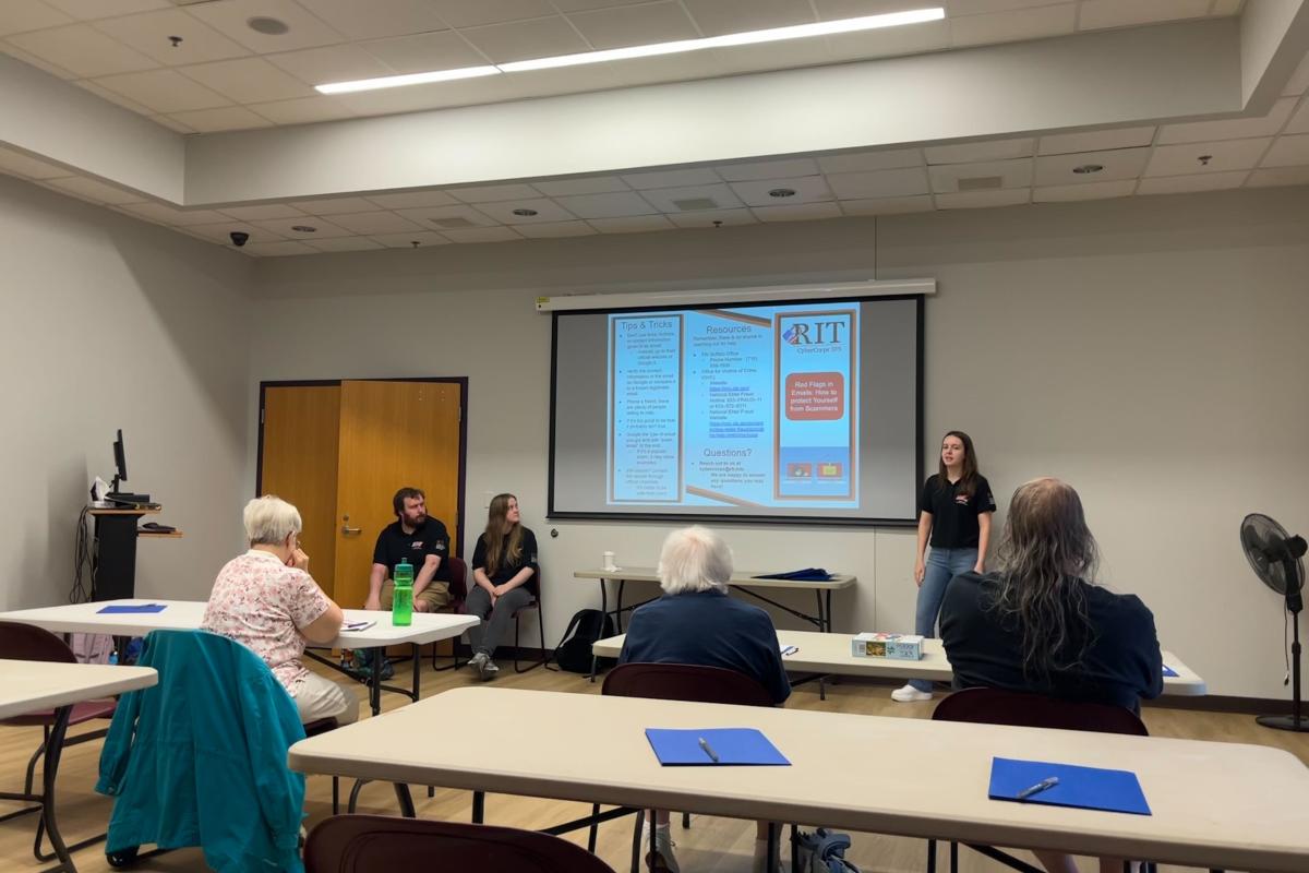Presenters lead a cybersecurity awareness workshop for community members in a classroom, with attendees seated at tables and educational slides displayed on a screen.