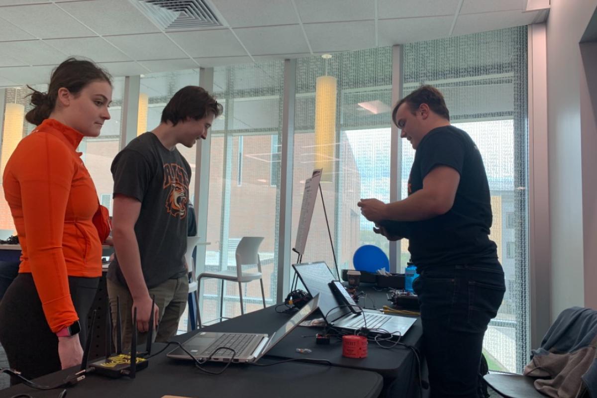 Attendees visit a technology demonstration table with laptops, networking equipment, and electronics in a bright event space with floor-to-ceiling windows.