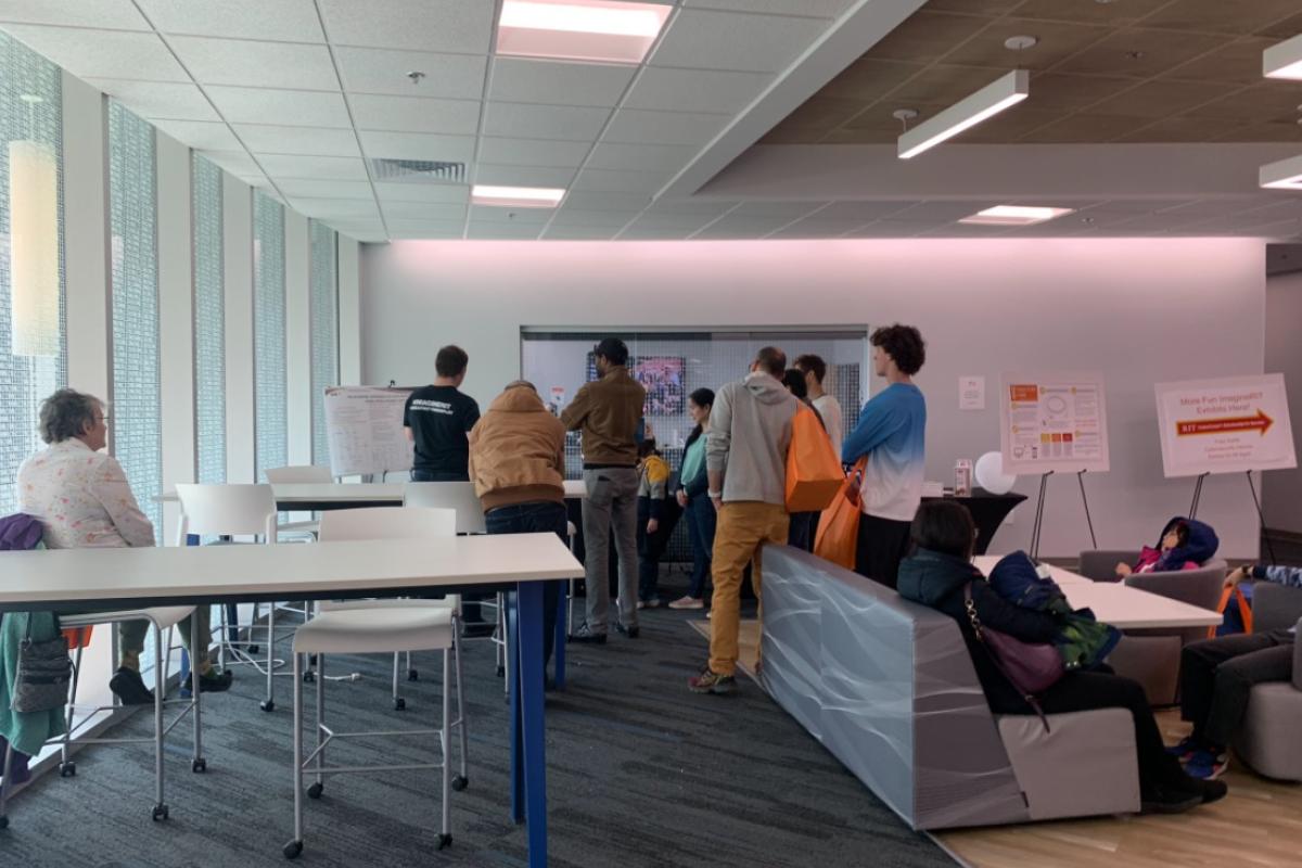Visitors gather around technology exhibits in a bright event space, viewing demonstrations and displays while others sit at tables and lounge seating nearby.