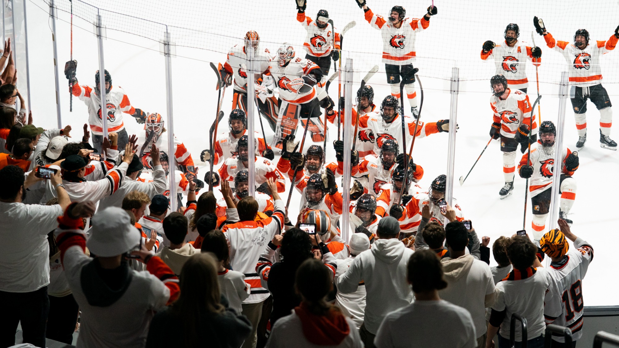 College hockey players in white and orange uniforms gather along the boards after a game, raising their sticks and celebrating with a crowd of fans reaching over the glass from the stands.
