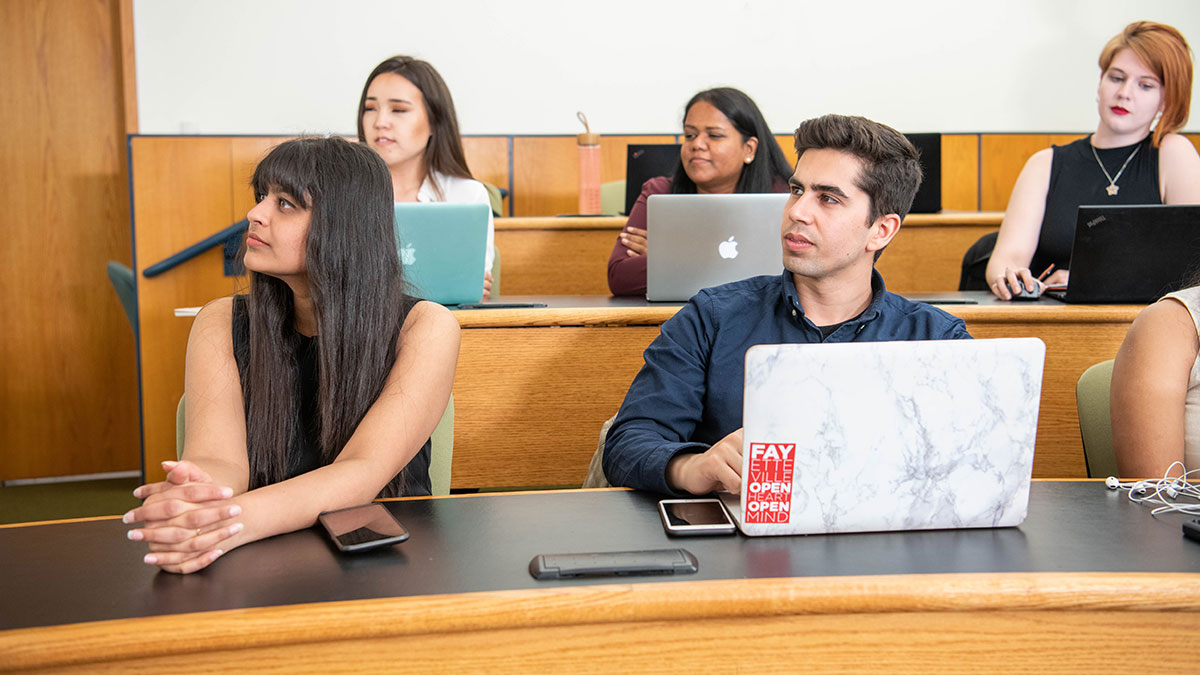 Students attentively listening during a classroom lecture, with open laptops in front of them.