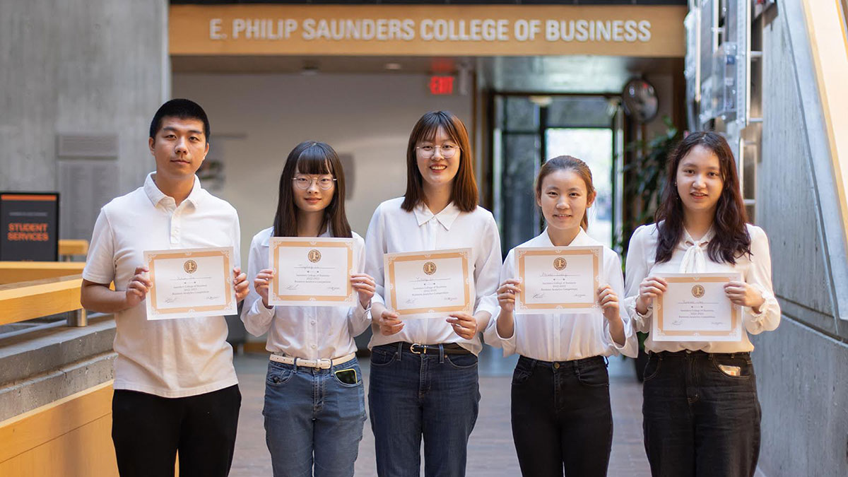 Five students holding certificates and posing for a group photo in a business school lobby.