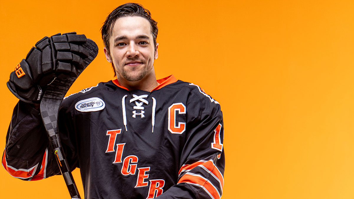 A hockey player wearing a Tigers jersey and holding a hockey stick with an orange background.