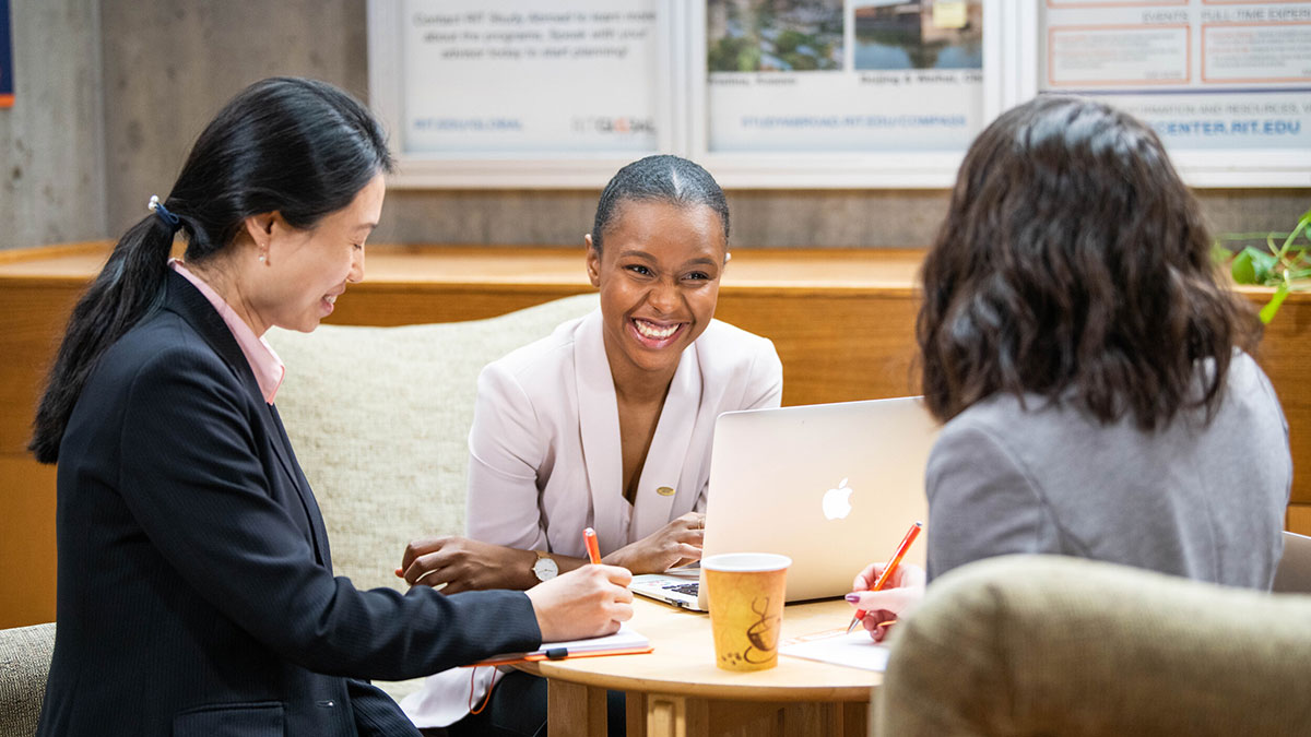 Three professional women smiling and collaborating on a project at a table with a laptop.
