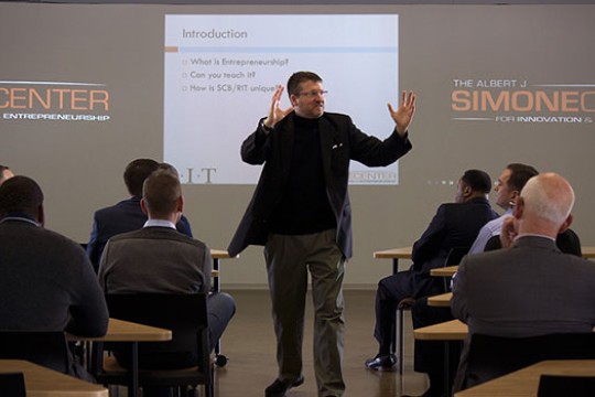 Richard DeMartino with hands up lecturing in front of people.