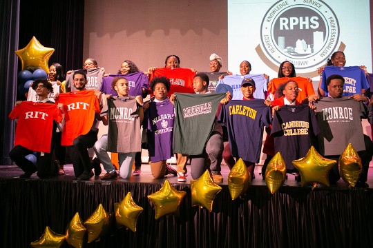 A group of graduating students from Rochester Prep High School stand on a stage and hold up t-shirts from the colleges they have been accepted into for the fall.