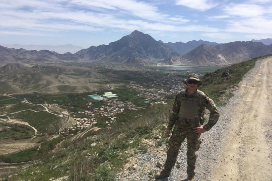 American soldier stands on roadside with mountains in background.
