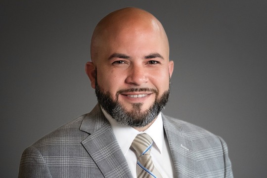 Orlando Ortiz sits for a headshot against a gray background wearing a gray plaid suit.