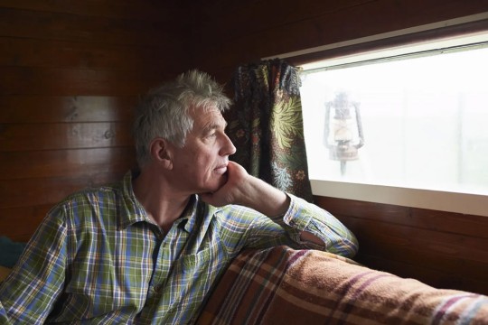 a man with gray hair and a plaid shirt looks out a window to a snowy landscape.
