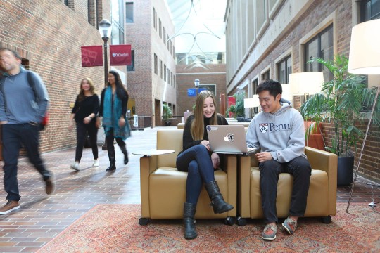 two students sit on chairs in the middle of a busy walkway between brick buildings.