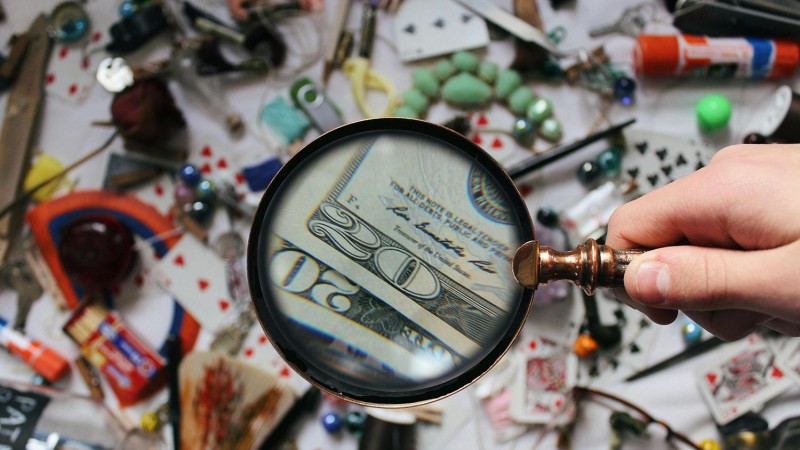 a cluttered desk with a person holding a magnifying glass to a U S 20 dollar bill.