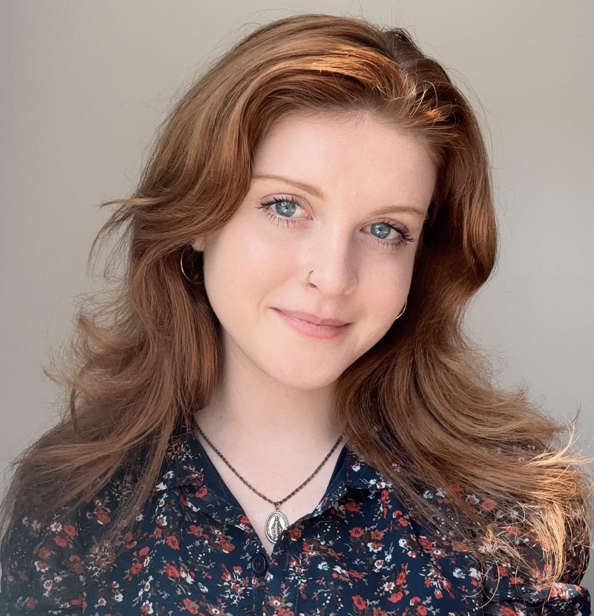 Headshot of Maya, a white woman with long auburn hair, in a floral navy blue top and smiling against a plain gray background. 