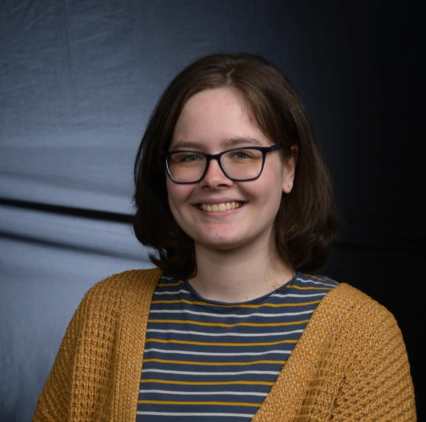 Headshot of Stephanie, a white woman with brown shoulder-length hair, blue eyes, and glasses. She is smiling and wears a blue striped shirt and a yellow cardigan.
