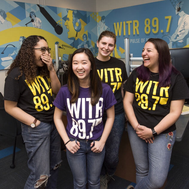 Four members of the W I T R Radio Station wearing W I T R shirts in their studio and laughing.