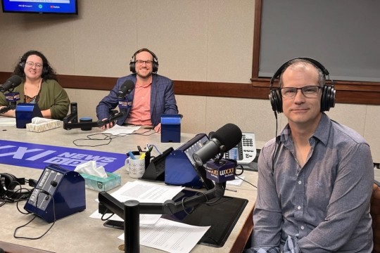 three people sit at a table wearing headphones and speaking in to microphones.