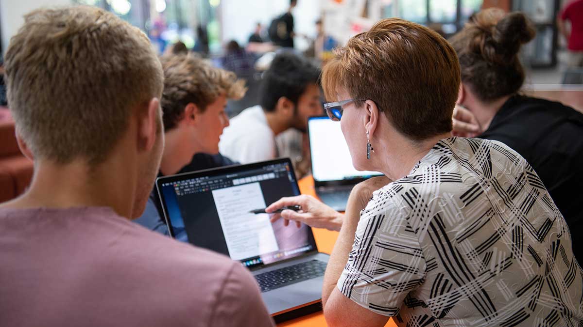 A woman points to information on a laptop screen while assisting a student with a document.