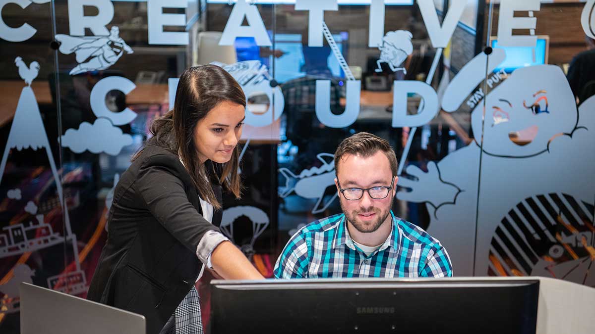 A woman points at a computer monitor while standing beside a man in an office decorated with Creative Cloud graphics.