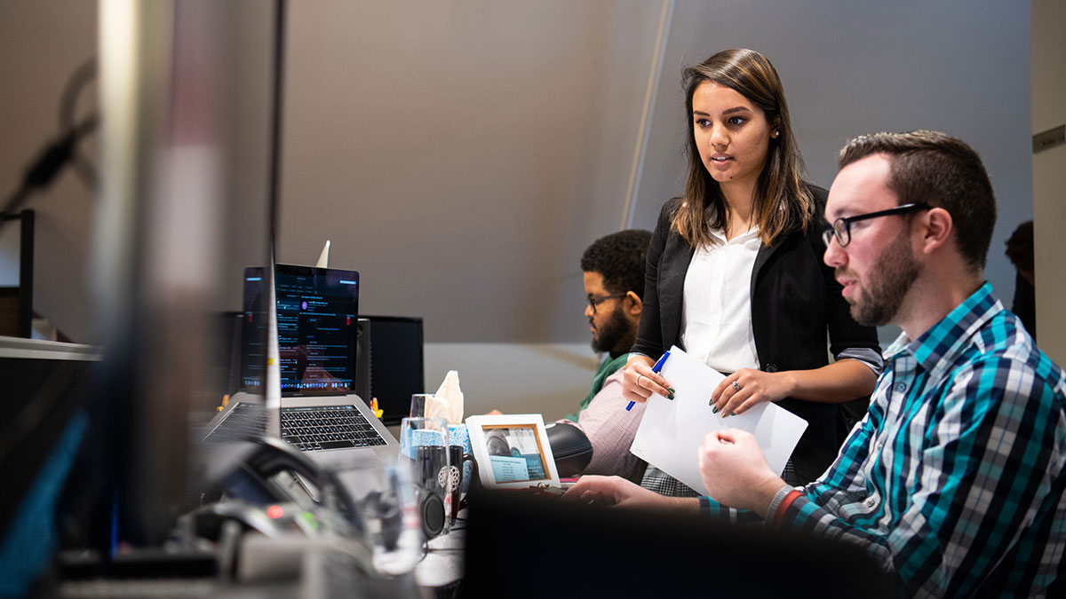 A woman stands beside a seated man discussing work in a modern office with computers.