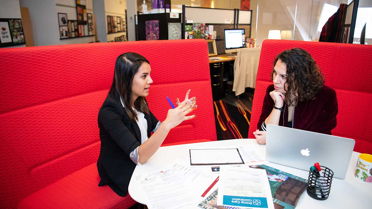 Two women sit in a red booth having a discussion with papers and a laptop on the table.