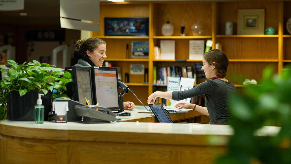 Two women smile and talk at a library service desk with computer stations and bookshelves in the background.