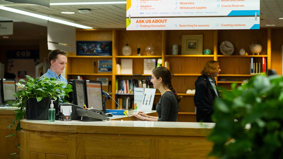 A smiling librarian assists a student at the library desk with computers and bookshelves in the background.