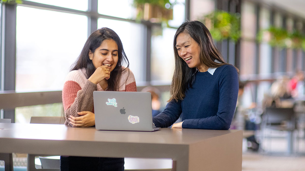 two students smiling while looking at a laptop together.