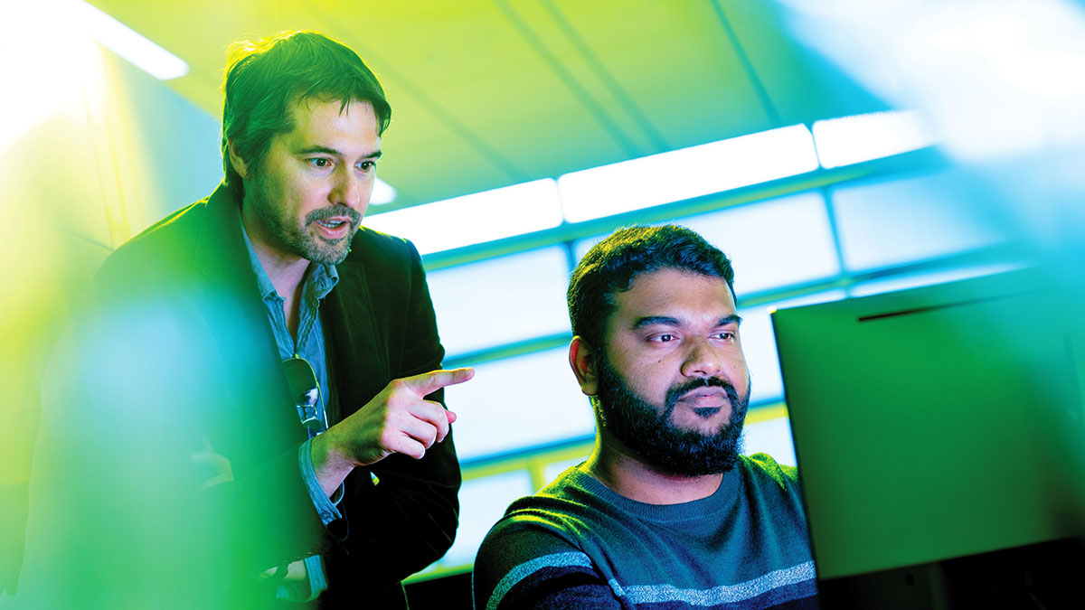 student seated at a computer with a professor standing behind him and pointing at the screen.