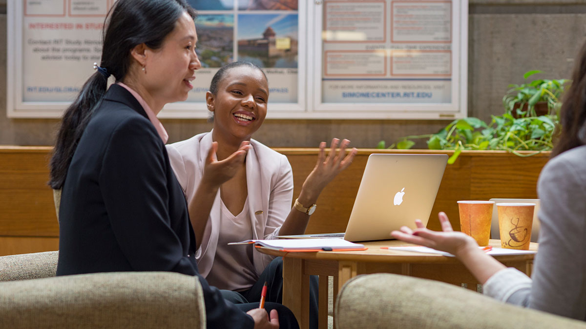 Three people in professional attire smile and talk around a table with a laptop.
