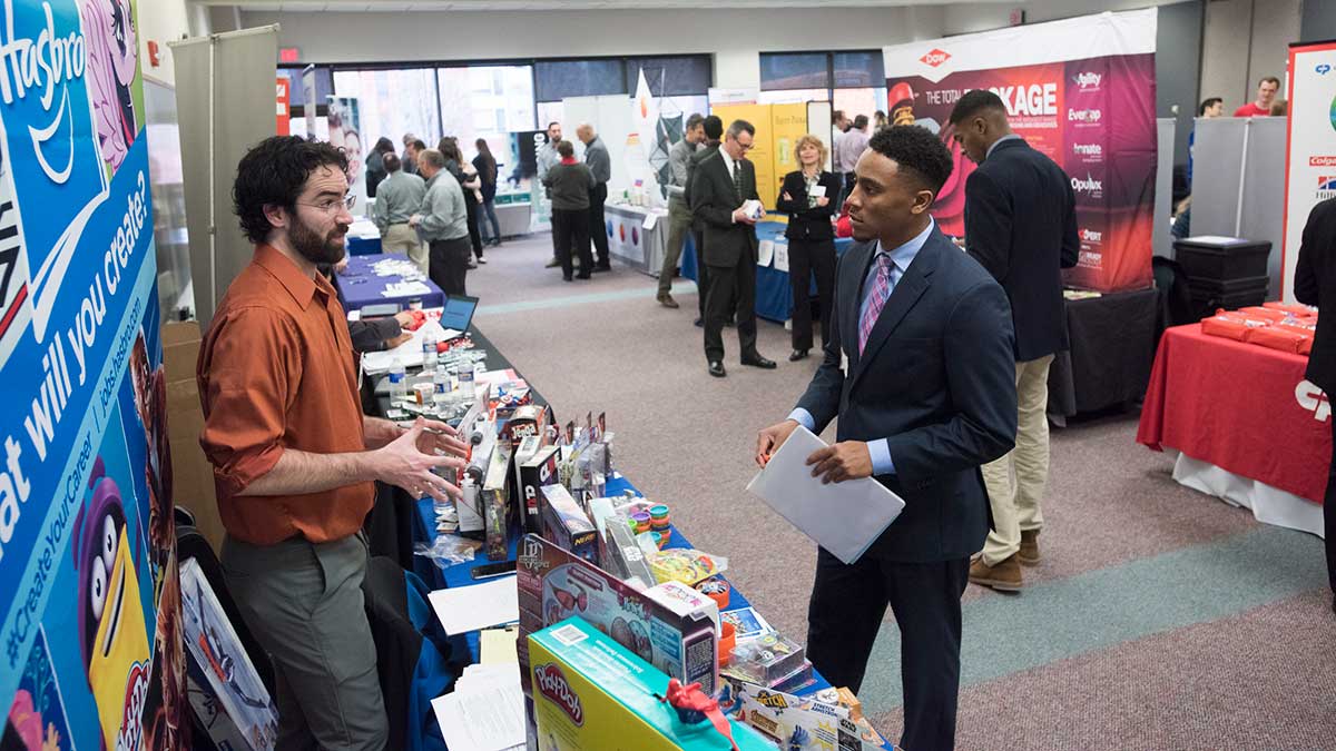 A job seeker in a suit speaks with a recruiter at a booth during a career fair.