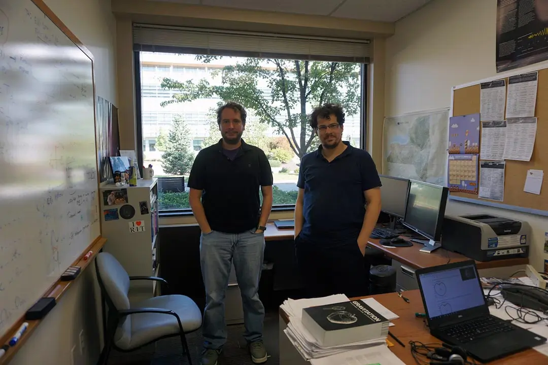 Two researchers stand in a small office space with a large window.