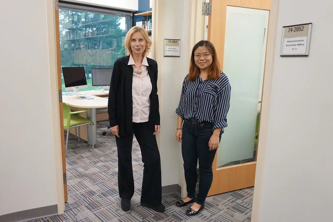 Two women stand outside their office doors in a brightly lit office area.