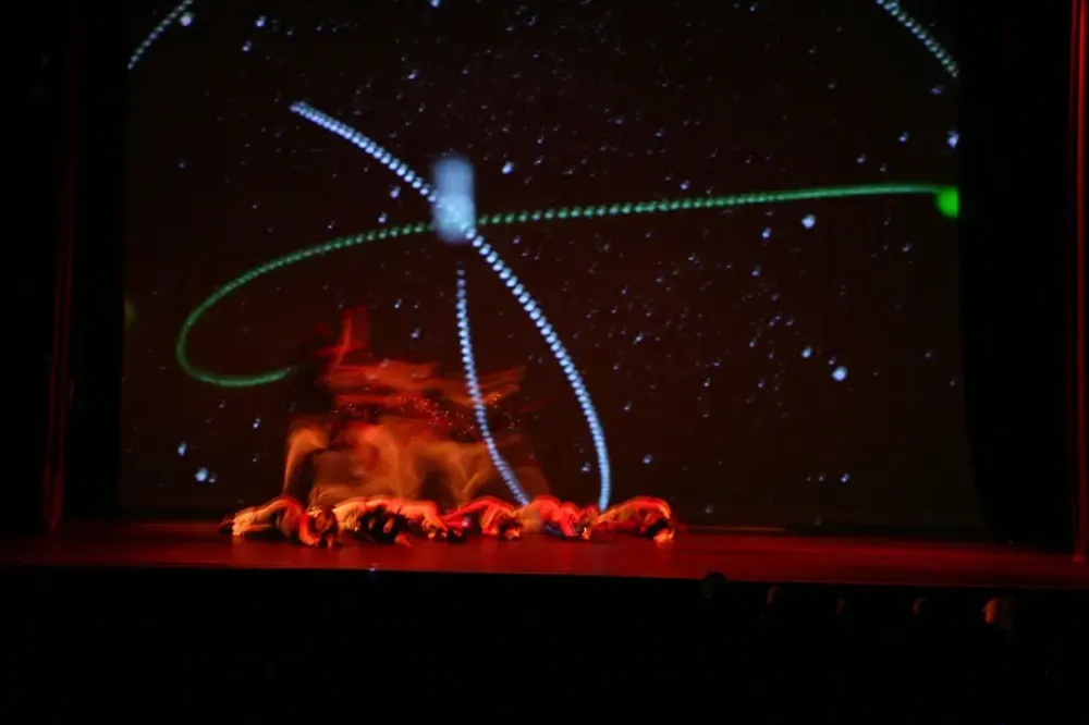 Dancers on a stage crouched down in a group with a red light shining on them, with images of space in the background.