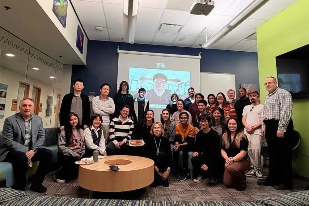 A large group of people poses for a photo in a lounge area, with a person displayed on a screen in the background.