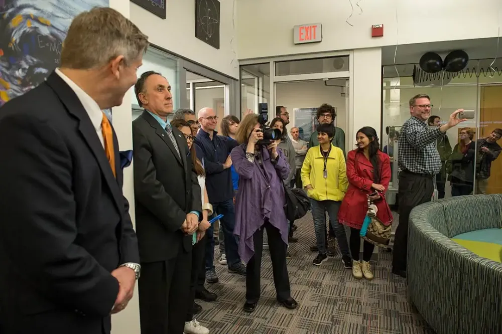 A photographer takes a photo during an event in which attendees stand along the back of a room.