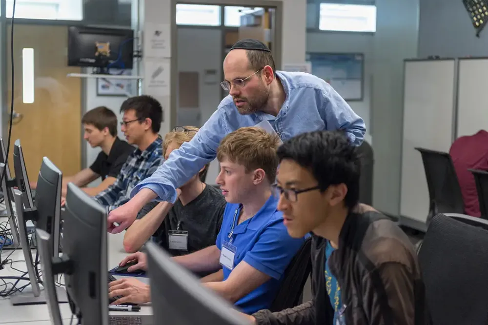 A professor helps a student by pointing to someone on his computer.