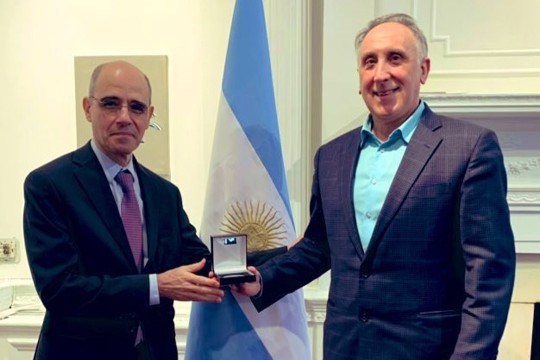 two older men stand next to each other with the man on the left presenting the other with a small box that contains an award. a blue and white flag with a yellow sun is behind them.