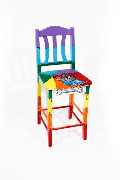 A dog with a large hanging tongue dressed in a blue cape and wizard hat with a large purple gem as the clasp and a light purple dog collar. The dog is holding up a wand tipped with a star in one hand and out of the wand is flowing all the colors of the rainbow. The rainbow colors begin with purple at the top of the chair and proceeds sequentially downwards ending with red. 