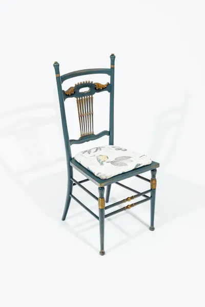 Classic slat-back designed chair painted blue gray, with brushed gold/bronze accents. The chair has a decorative crest rail with gold painted winged motifs. There is an upholstered seat, the fabric is a cream background printed with a vintage botanical/ zoology collage of birds, leaves branches and faux vintage script.