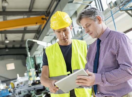 two men looking at tablet with factory floor in the background